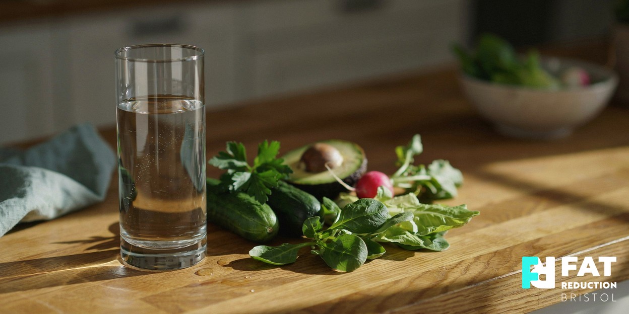 A still life of a glass of water and fresh green vegetables on a warm wooden kitchen surface in soft daylight