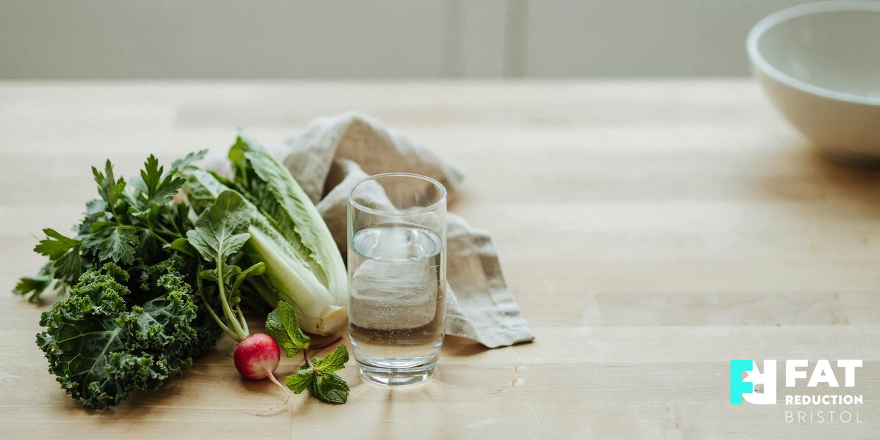 A photographic still-life of fresh green vegetables and a glass of water arranged on a pale wooden kitchen surface in soft natural daylight