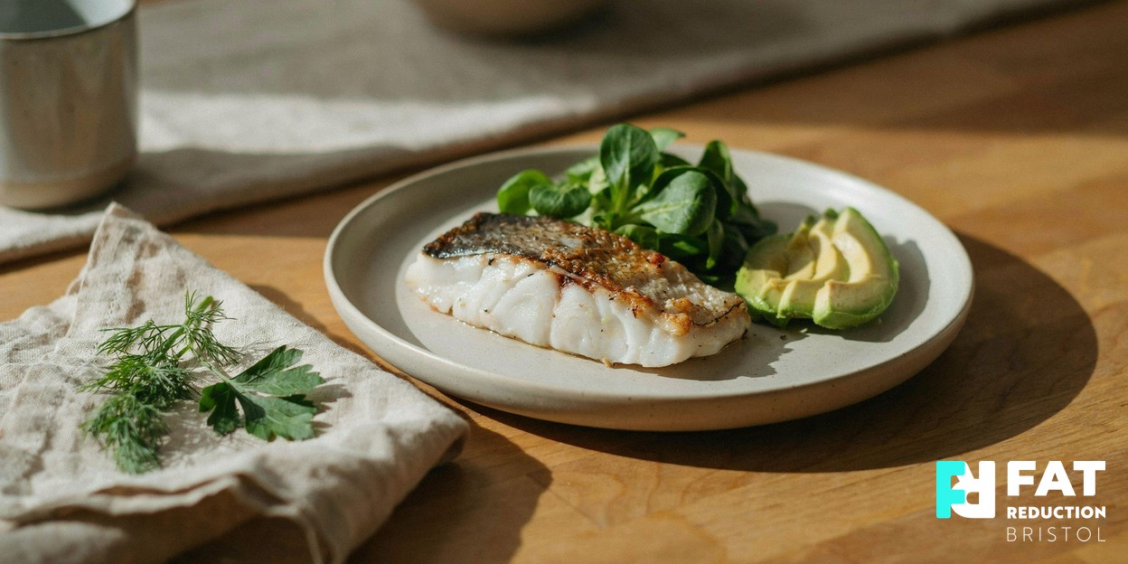 A colourful protein-rich healthy meal of grilled fish, leafy greens and avocado arranged on a pale ceramic plate on a warm wooden table in soft daylight