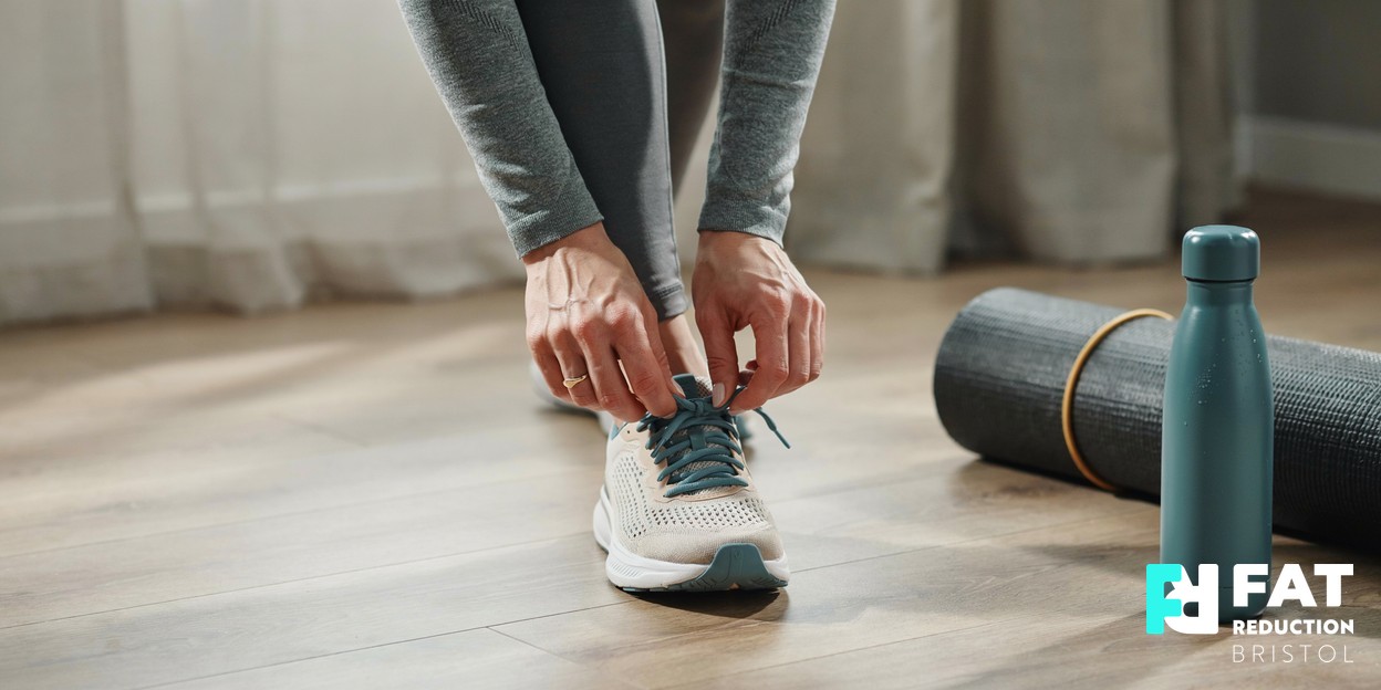 A woman’s hands lacing up a running shoe beside a rolled exercise mat on a light wooden floor in soft morning daylight