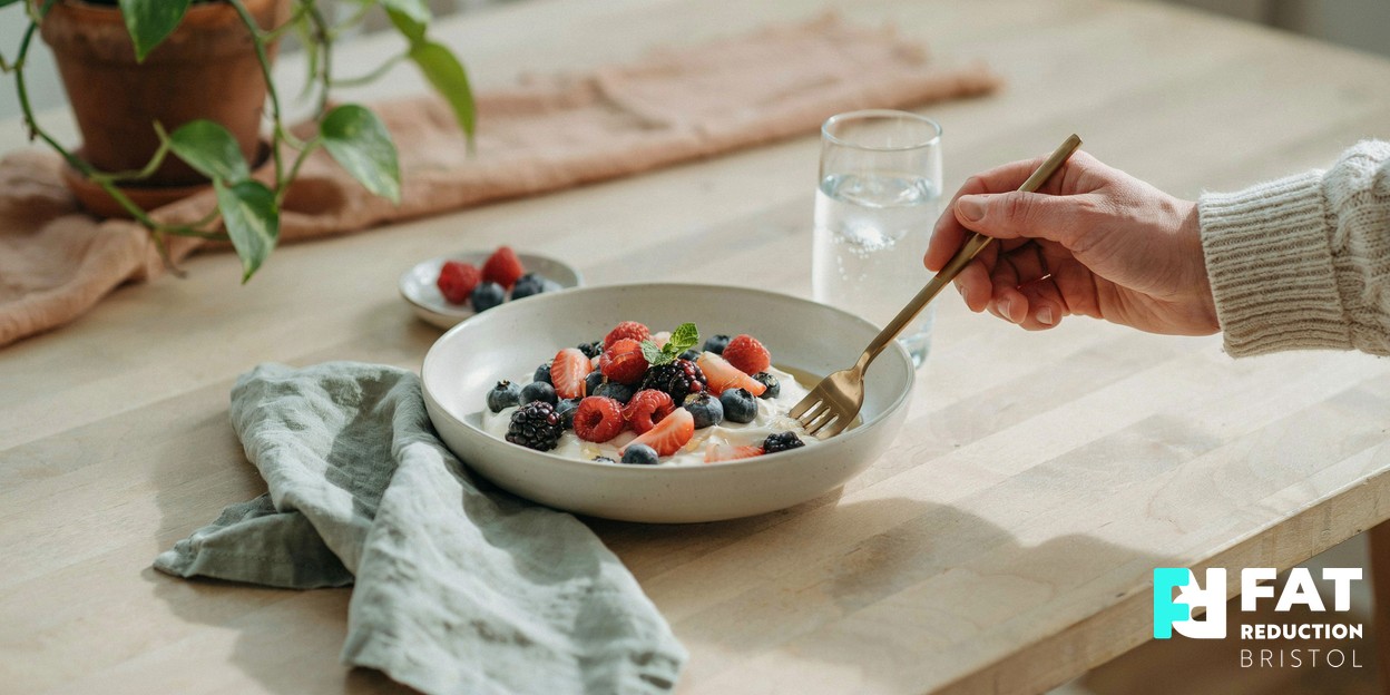 A healthy breakfast of fresh berries, natural yoghurt and a glass of water on a sunlit wooden table, with a hand resting a fork at the edge of the plate