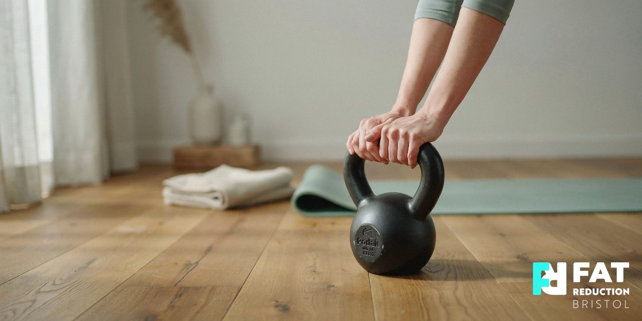 A photograph of a woman’s hands resting on a kettlebell on a wooden floor in a bright, calm home gym