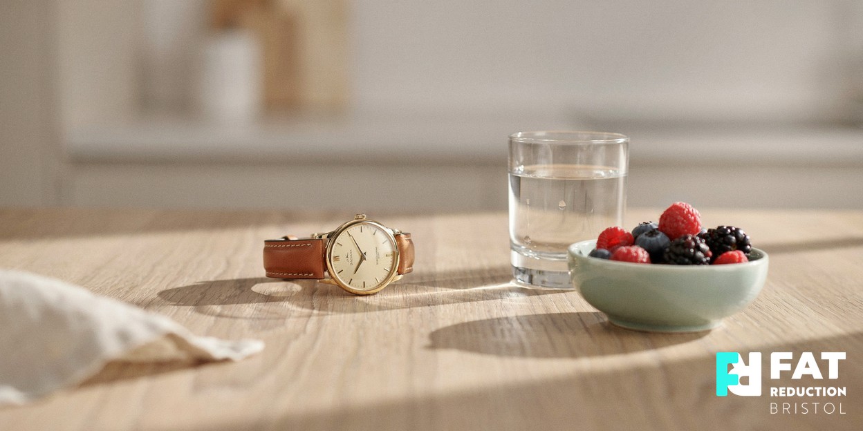 A still-life photograph of a classic wristwatch resting beside a clear glass of water and a small ceramic bowl of berries on a pale wooden table in soft daylight
