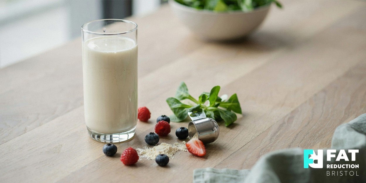 A still-life photograph of a pale meal-replacement shake in a glass on a light wooden table beside fresh whole foods — leafy greens, berries and a measuring scoop — in soft natural daylight