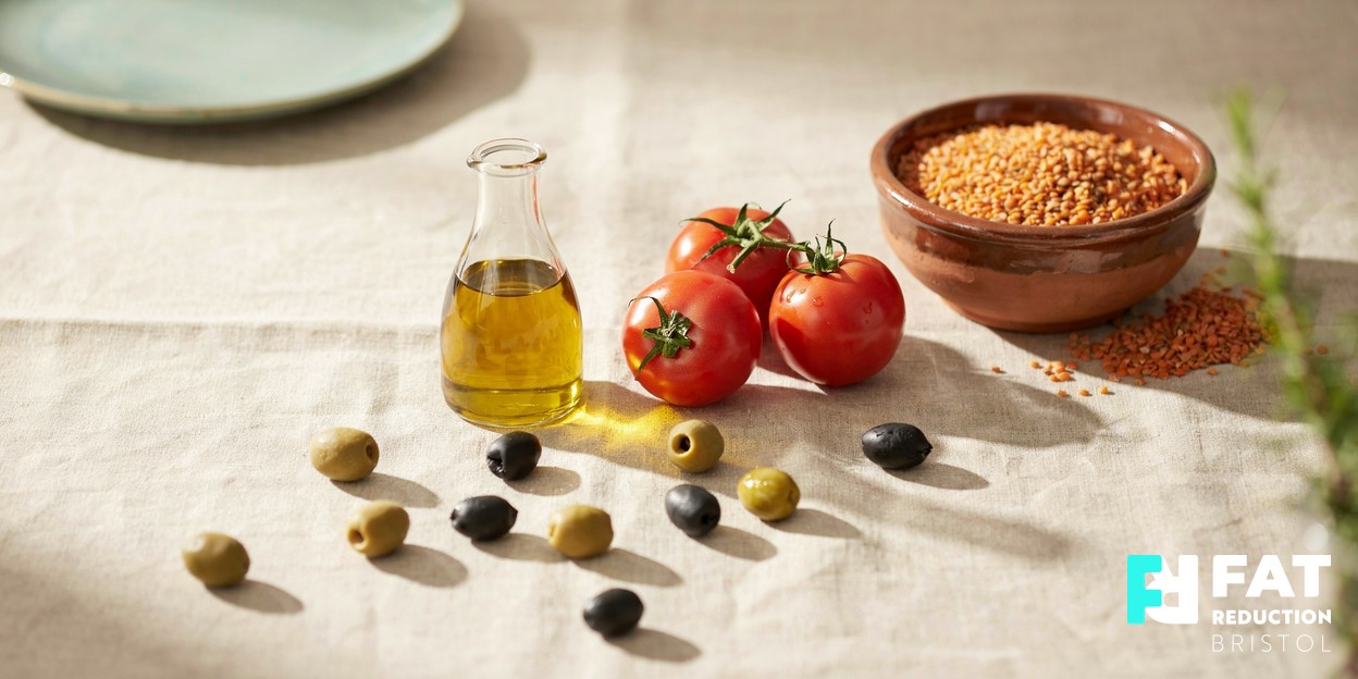 A still-life of Mediterranean staples on a warm neutral surface — a small jug of olive oil, ripe tomatoes, a bowl of dried lentils and scattered olives in soft daylight