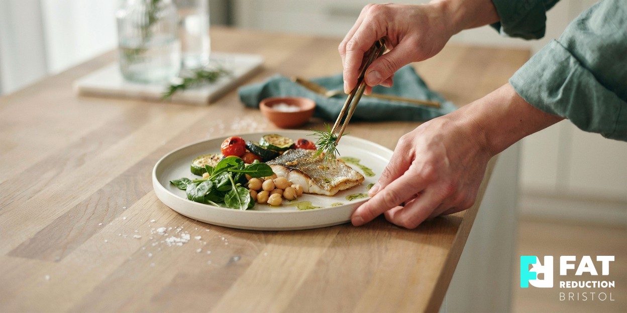 A person’s hands plating a fresh Mediterranean meal of leafy greens, chickpeas and grilled fish on a ceramic plate at a bright table