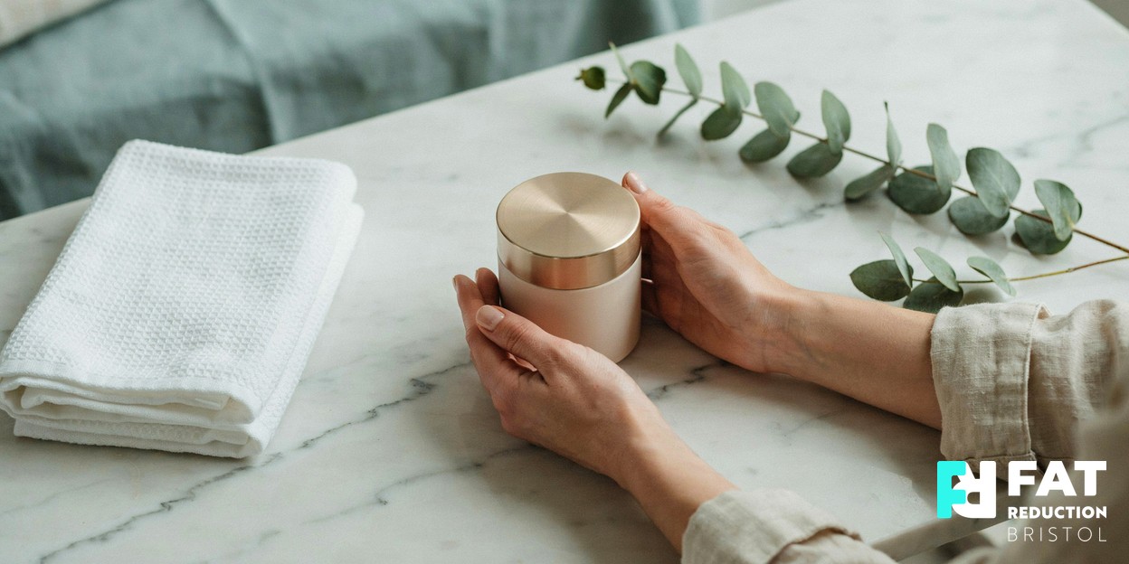 A woman’s hands cradling a ceramic skincare jar beside a folded towel and a sprig of eucalyptus on a calm marble surface