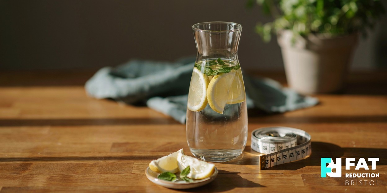 A coiled cloth tape measure resting beside a glass carafe of water with lemon and fresh mint on a warm wooden surface, in soft natural daylight
