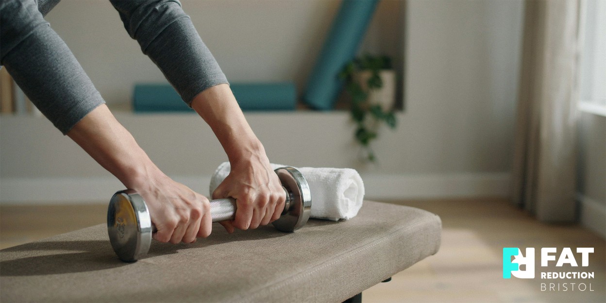 A person’s hands gripping a light dumbbell resting on a bench beside a rolled towel in a calm, bright home gym